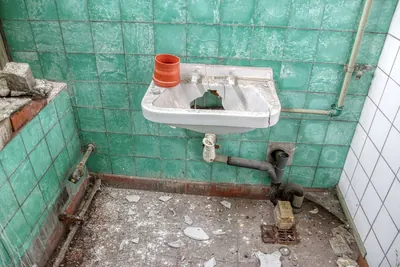 Damaged bathroom with a broken sink, exposed pipes and peeling tile